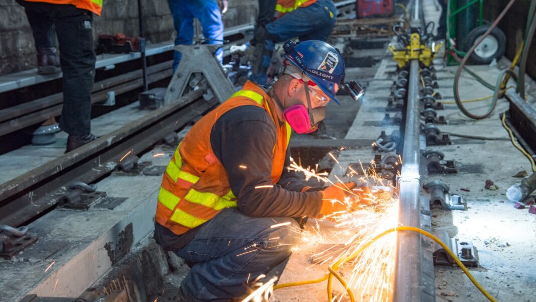 A Smaller Labor Force Complicates the Meaning of Every New Jobs Number A construction worker in a hard hat and safety vest grinds a steel rail, sending up sparks