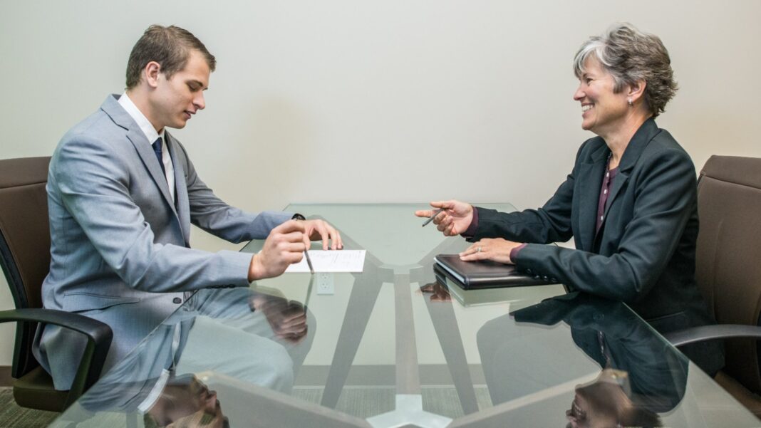 Advertised Pay Is Rising Unevenly Across Occupations as Job Postings Improve A young man in a suit and an older woman sit across a glass table during a job interview