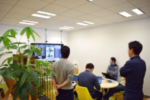 Office workers in Japan gather around a television screen in a bright office