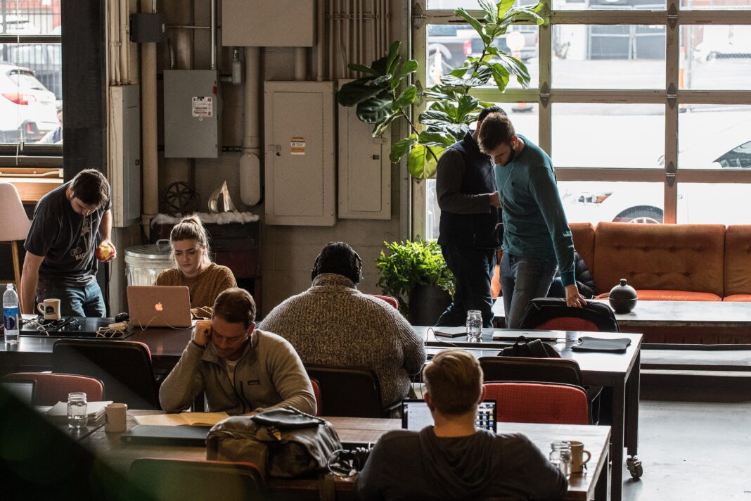 Most AI-Using Firms Have Not Redesigned Work Around It. That May Be the Harder Step People work on laptops at shared tables in a coworking office with large windows
