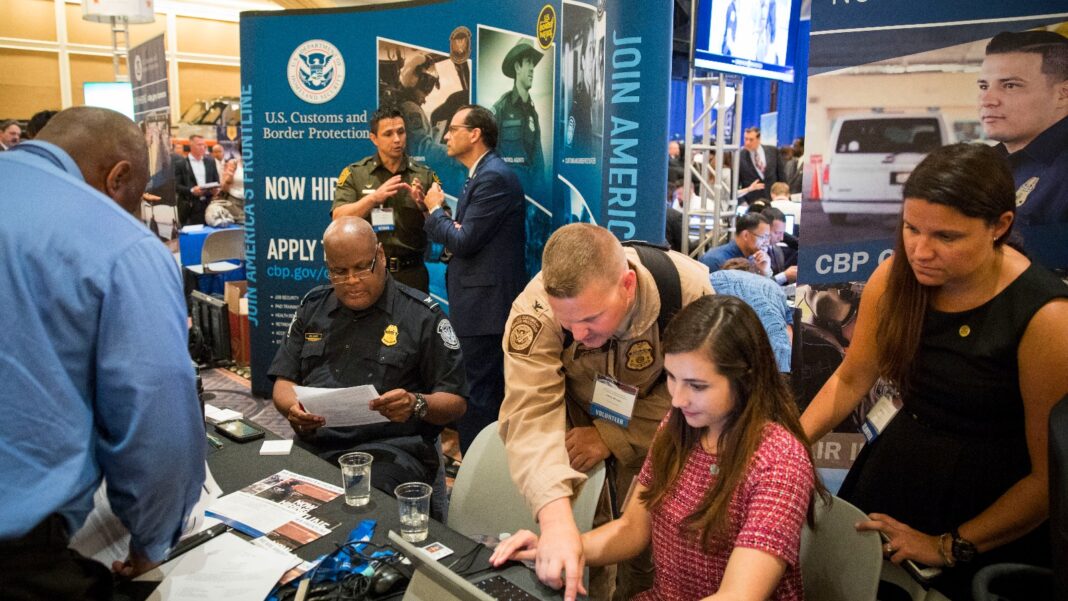 Uniformed recruiters and job seekers gather around a table at a crowded hiring-fair booth with a Now Hiring banner