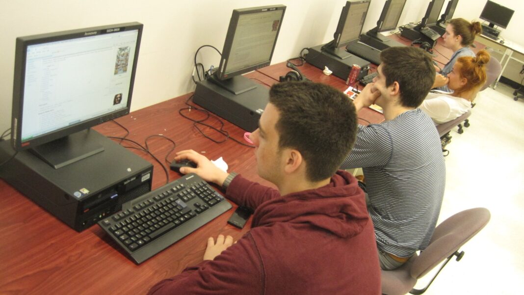 Young adults sit at desktop computers in a row along a wooden counter in a computer lab