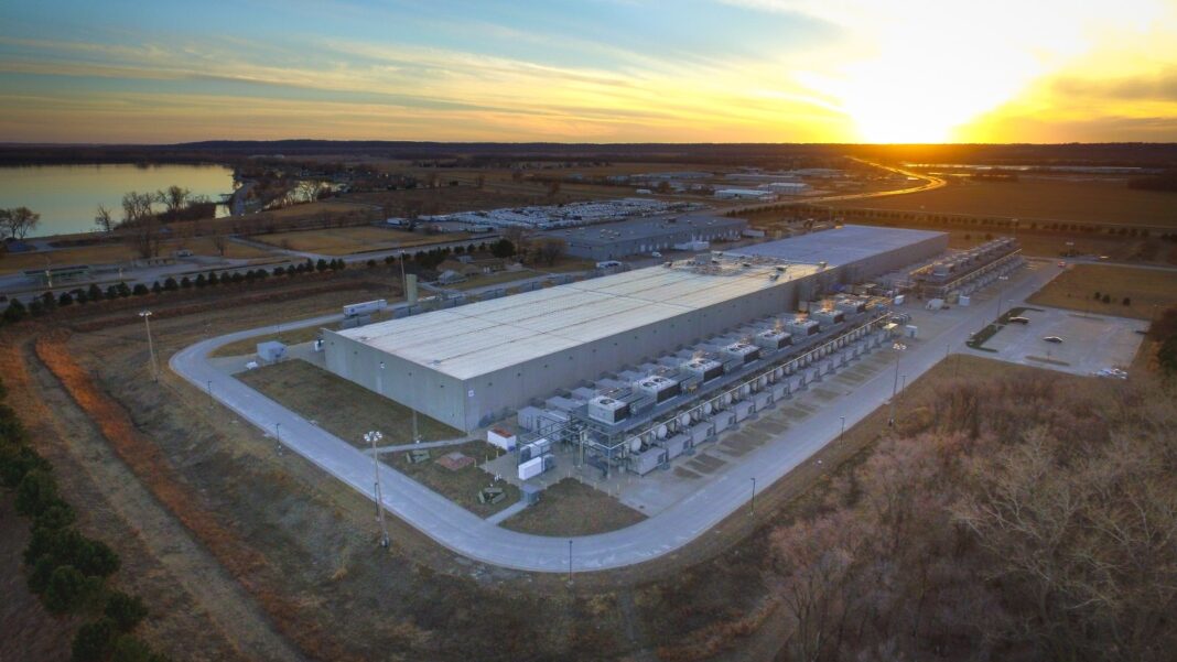 Data Centers Can Raise Local Payrolls and Living Costs at the Same Time, New Research Finds Aerial view at sunset of a large, low data-center building surrounded by cooling equipment and open land