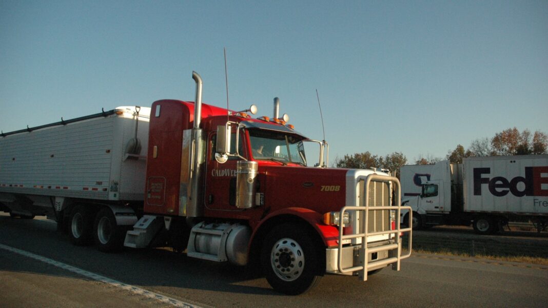 Federal Suit Challenges Age-Based Screening Rules for Texas Trucking Applicants A red semi truck hauling a trailer stands at a highway truck stop at dusk with another truck behind it