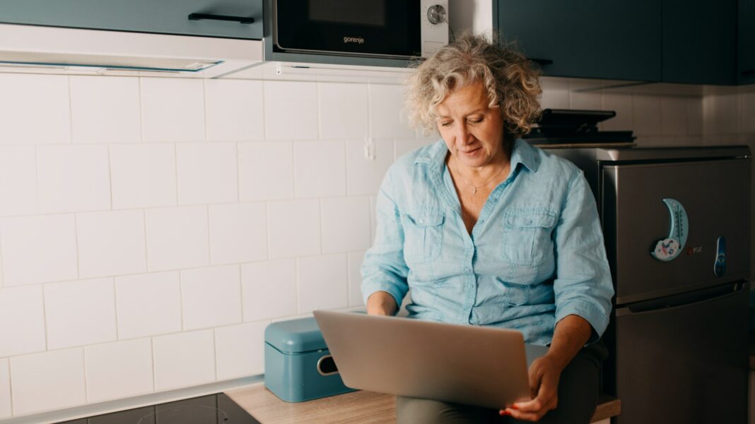 Flexible Hours and Work-from-Home Are Different Benefits. New Federal Data Show Why A woman in a denim shirt works on a laptop while sitting in a modern white kitchen