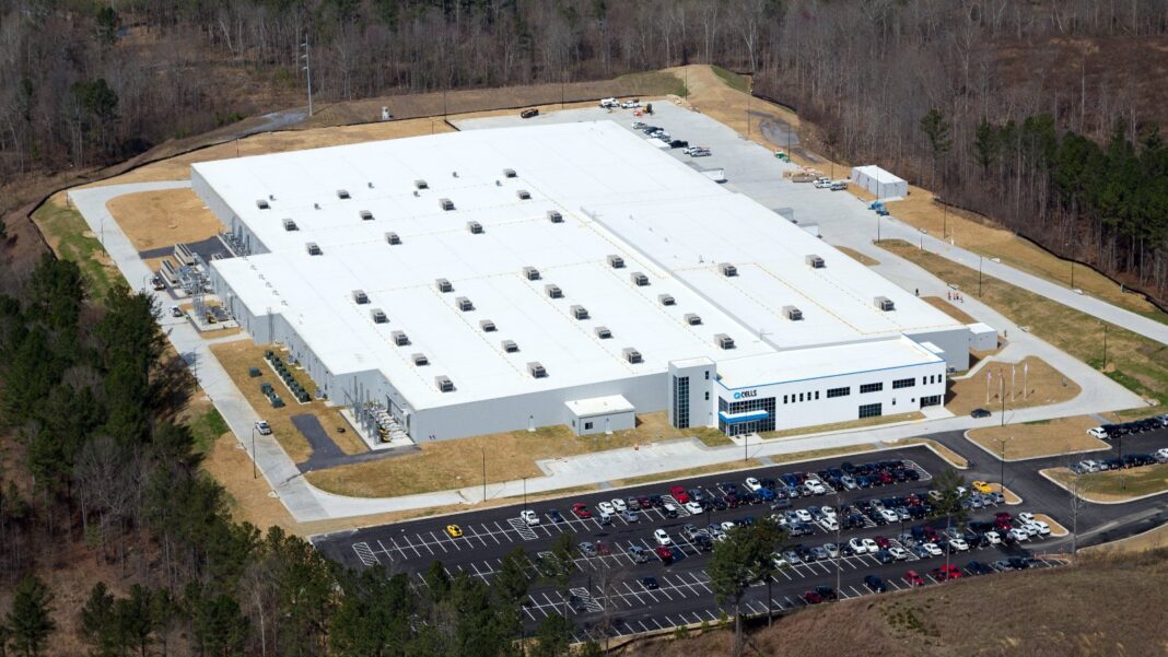 Aerial view of a large white factory building with a full parking lot surrounded by wooded hills