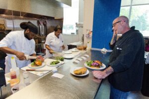 A chef reads lunch orders to two line cooks at a restaurant kitchen counter