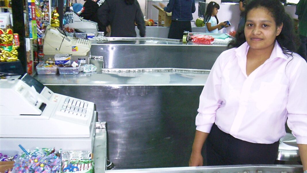 A cashier in a white blouse stands beside a checkout register in a grocery store