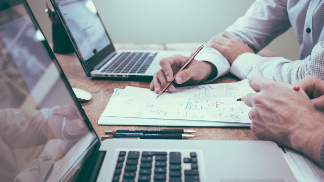 PwC Survey Finds AI Use Rising While Access to Workplace Learning Falls Close-up of two people at a desk with open laptops, one writing on a sheet of paper with a pencil