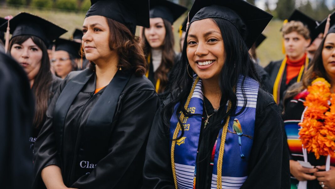 Smiling graduates in black caps and gowns, one wearing a colorful stole, gather at an outdoor commencement ceremony