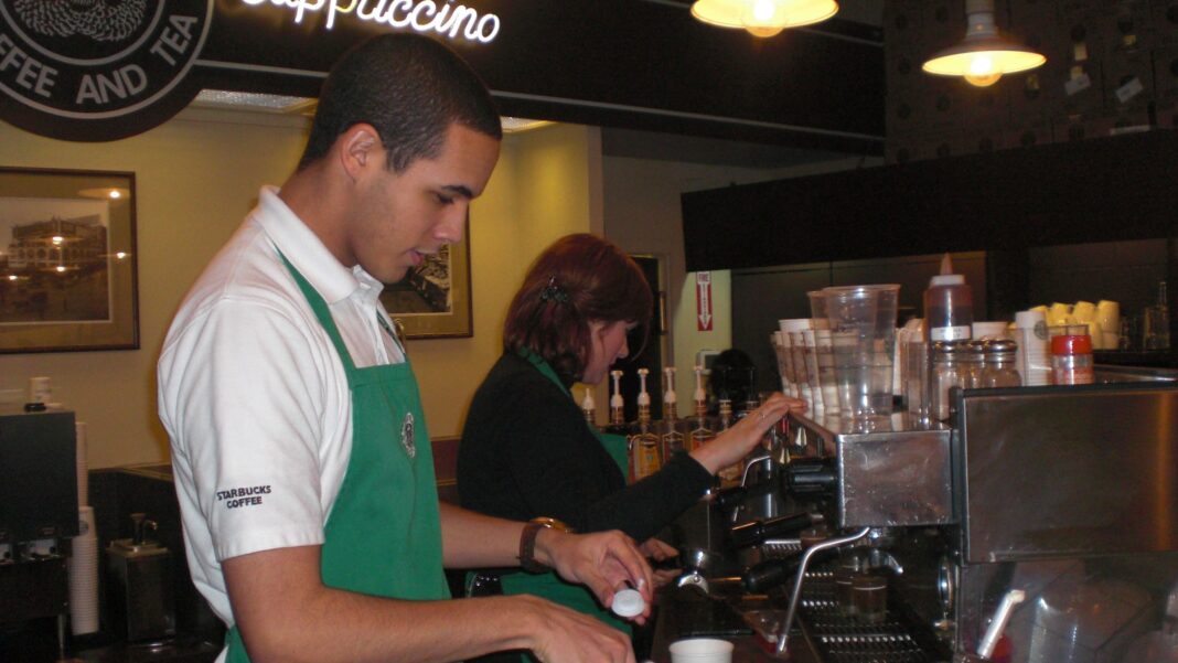 Starbucks Is Closing About 250 North America Stores While Building a New Technology Hub in India Two Starbucks baristas in green aprons prepare drinks behind the counter of a small coffee shop