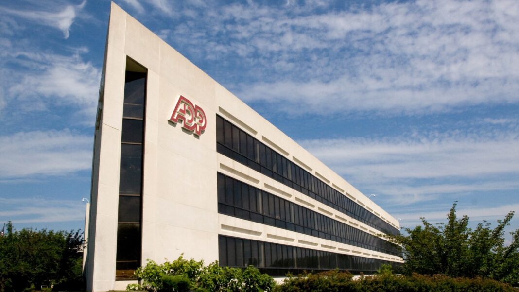 ADP’s Weekly Hiring Gauge Eased in Late September After Five Weeks of Gains A long, angular white office building with dark ribbon windows and a red ADP logo, behind a lawn and shrubs under a blue sky with clouds