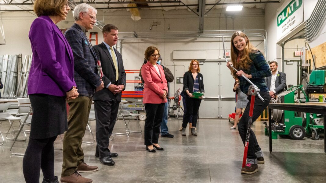 Amazon Plans to Train Up to 100,000 Workers a Year for Data-Center-Era Skilled Jobs A young woman demonstrates a long-handled conduit bending tool on a concrete floor as a group of visitors in business clothes watches in a training workshop