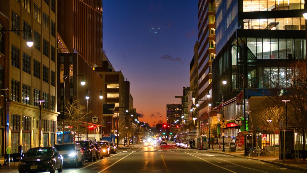 AstraZeneca Says $1 Billion-Plus Massachusetts Investment Will Expand Its Workforce There by More Than 50% A city street at dusk lined with modern glass office and laboratory buildings, with car headlights and an orange sky in the distance