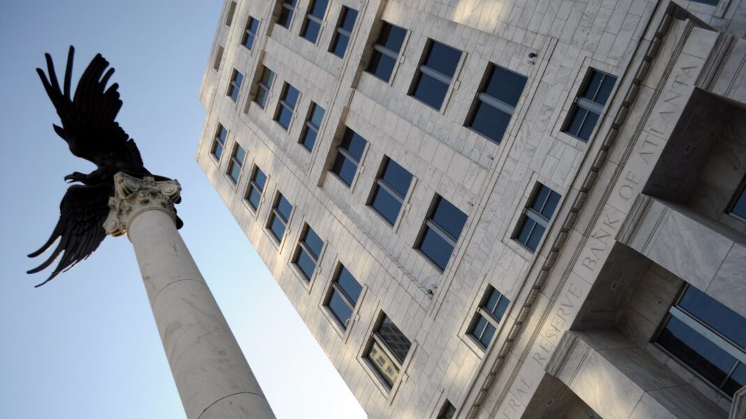 Low-angle view of a pale stone office tower carved with the words Federal Reserve Bank of Atlanta, beside a tall white column topped by a black eagle sculpture against a blue sky.
