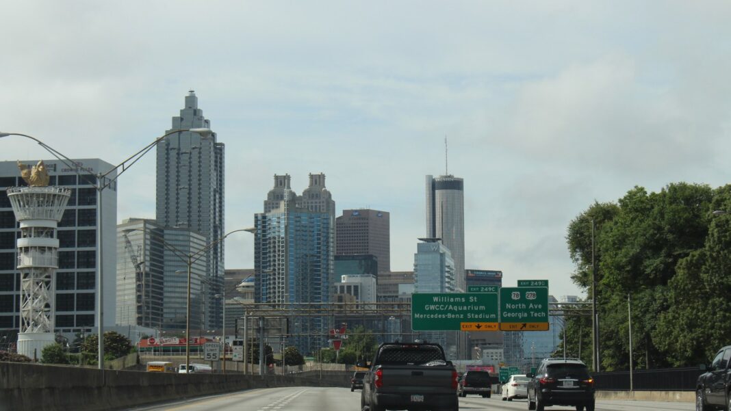 The Stitch Shows New Phase One Designs; Street Work Comes First, the Highway Cap Waits on Funding Traffic on a multilane interstate heading toward the downtown Atlanta skyline under green overhead exit signs on a cloudy day