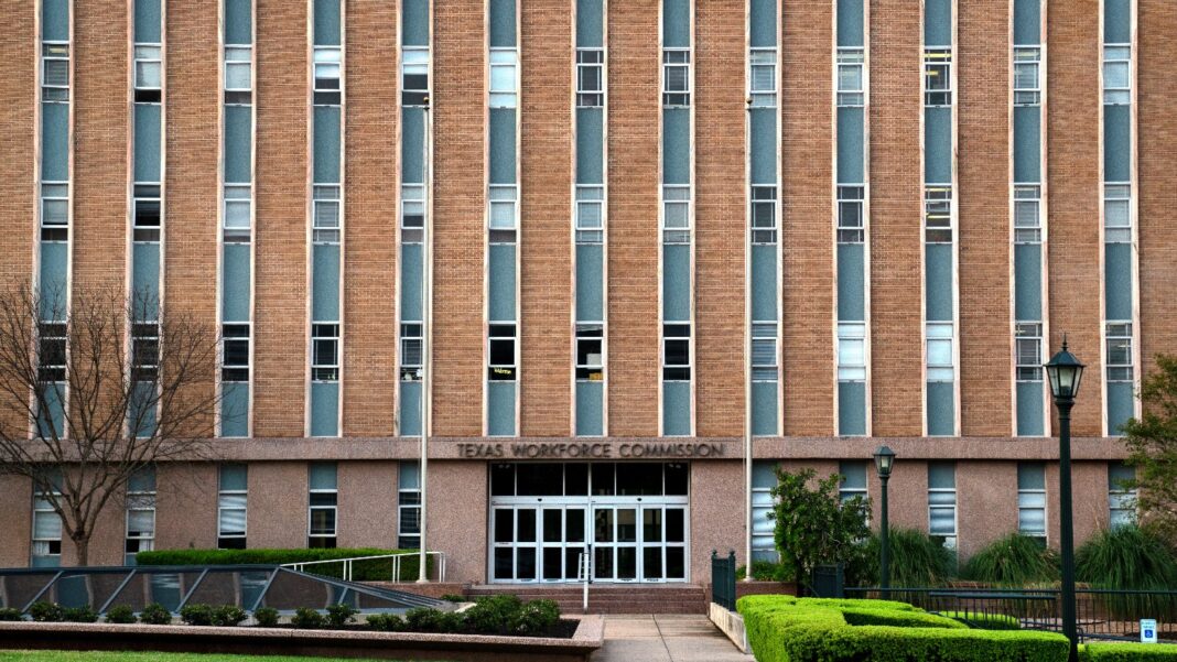 Black Unemployment Rose to 7.0% in September as More Workers Entered the Labor Force A tall tan-brick office building with narrow vertical windows and a glass entrance under a sign reading Texas Workforce Commission, framed by hedges and a bare tree