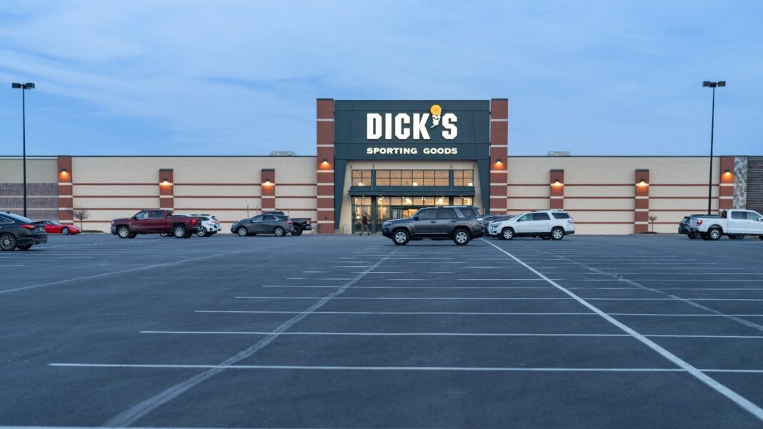 DICK’S and Foot Locker Plan More Than 11,000 Holiday Hires, Down From About 14,000 Last Year The front of a DICK'S Sporting Goods store with a lit sign over the entrance, seen across a large parking lot with several parked pickup trucks and SUVs at dusk.