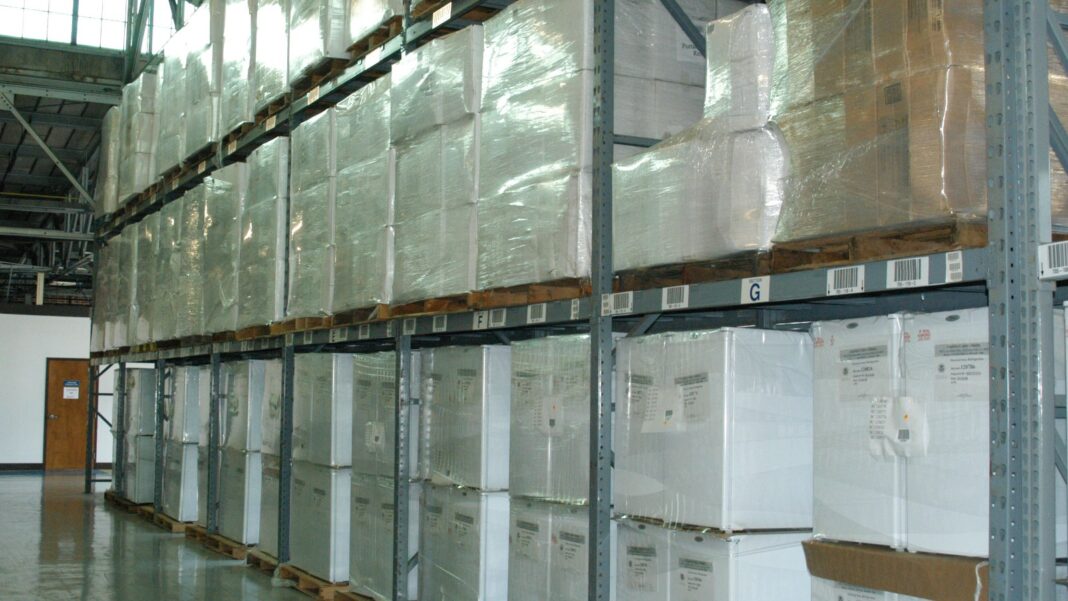Long rows of tall metal warehouse racks stacked with white boxed goods on pallets along a polished concrete aisle