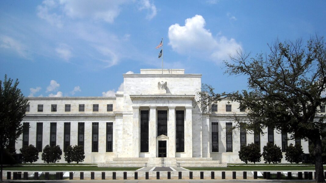 Fed’s October Beige Book: Hiring Mostly Flat Nationwide as Price Pressures Build A white marble neoclassical building with tall columns and an American flag on its roof, set behind a lawn and trees under a blue sky with clouds.