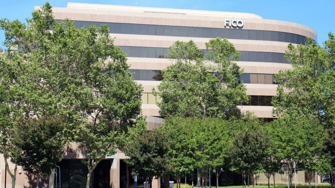 FICO Plans to Eliminate About 15% of Positions in a Second Restructuring in a Year A curved, multistory beige office building with a FICO sign near the roof, partly hidden behind green trees under a blue sky