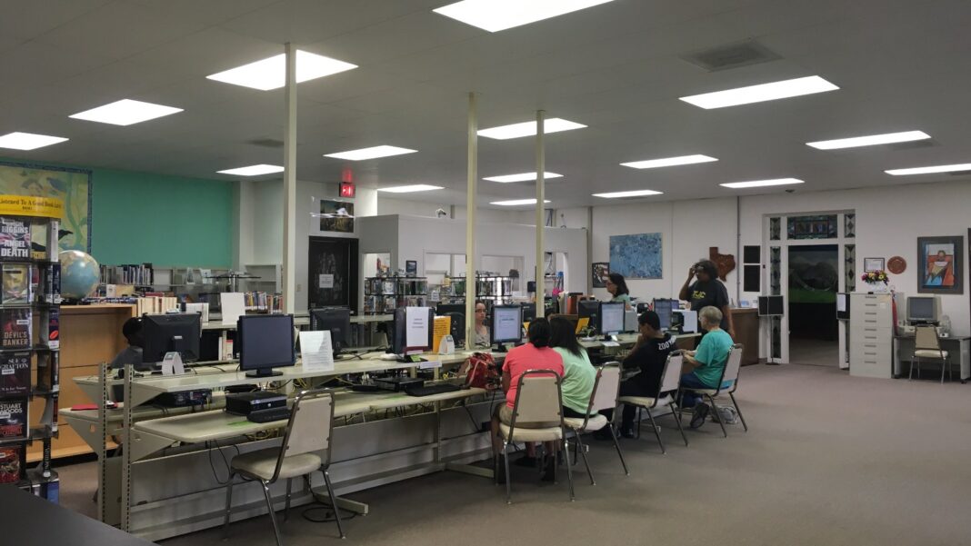 Adults sit at rows of desktop computers in a brightly lit public-library computer lab