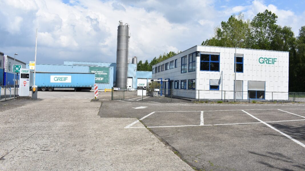 Greif Will Close Its Austell Paperboard Mill, Affecting About 90 Workers A white industrial office building with a green Greif logo beside a parking lot, with a blue warehouse and a tall silo behind it