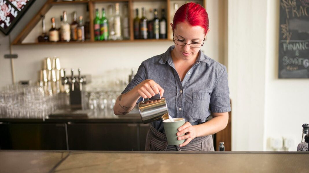 A barista with bright red hair pours steamed milk into a cup behind a long bar counter with bottles on shelves behind her