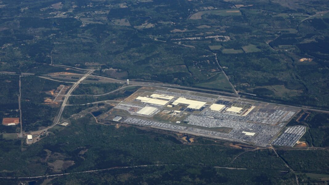 Aerial view of a large automotive assembly plant with rows of parked vehicles, surrounded by forest