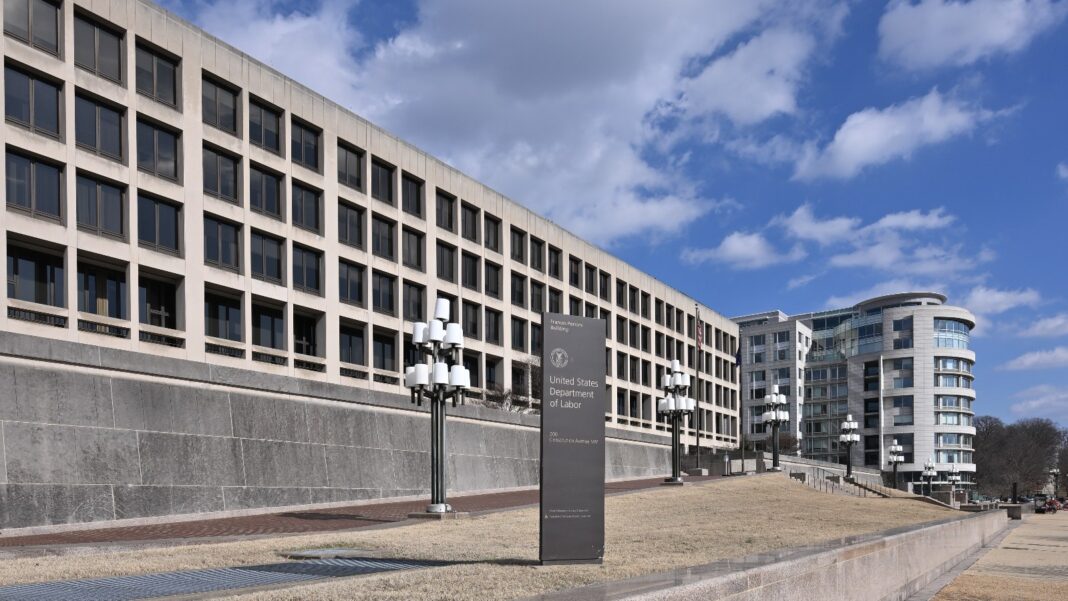 A dark sign reading United States Department of Labor stands on a plaza in front of a long, multistory concrete office building under a partly cloudy sky.