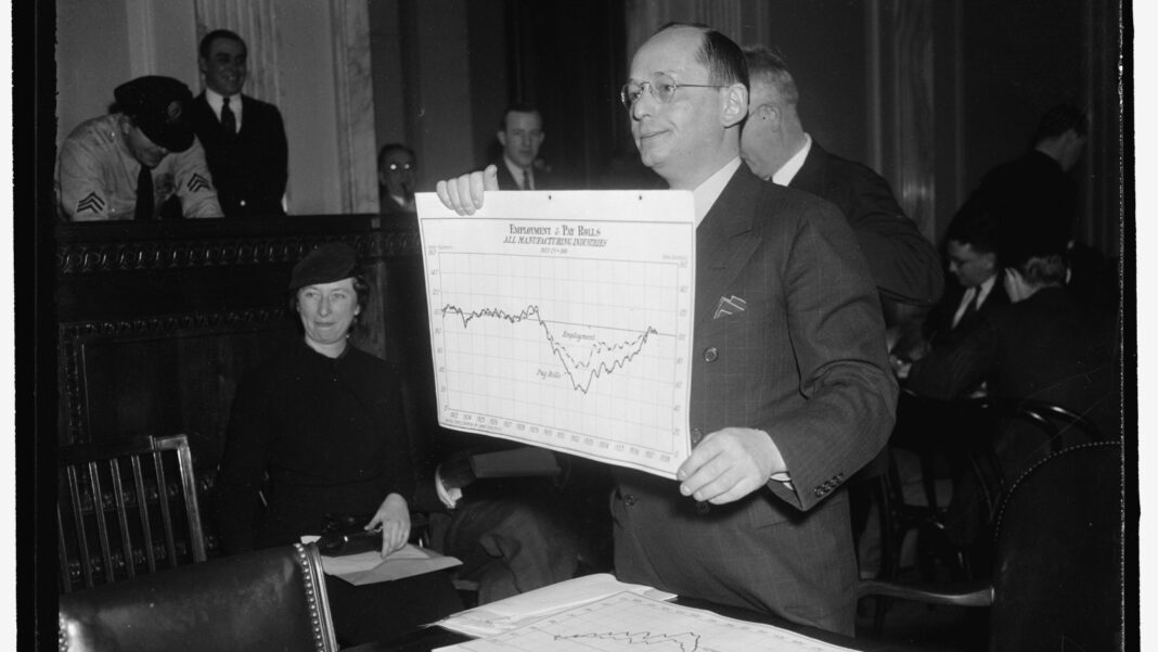 Black-and-white photo of a man in a suit holding up a line chart of employment before a committee, with other people seated behind him