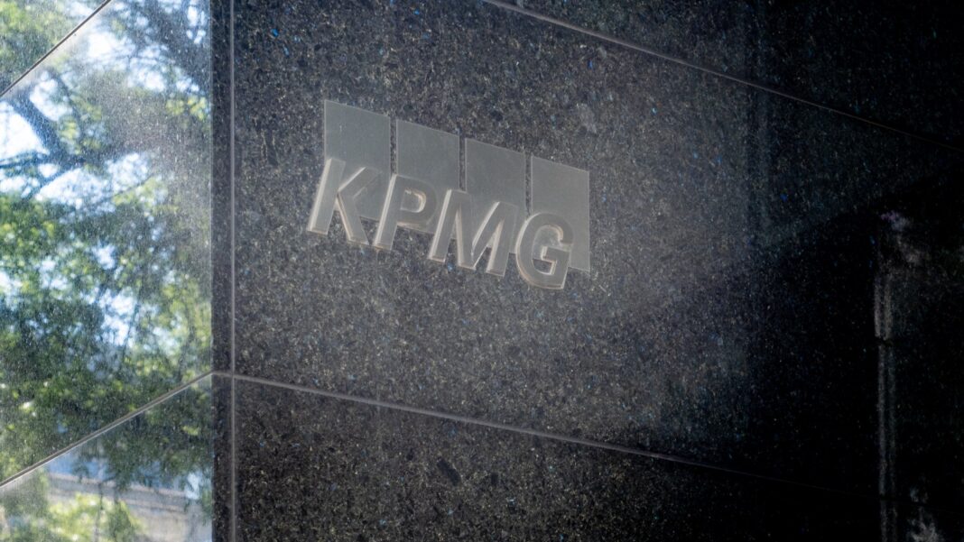 Brushed-metal KPMG logo mounted on a dark polished stone wall beside a glass window reflecting trees