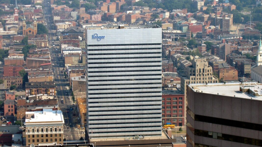 Kroger Plans to Cut About 3,000 Corporate Jobs Over Three Years Aerial view of a tall white office tower with the Kroger logo at the top, surrounded by brick buildings and a city street grid.