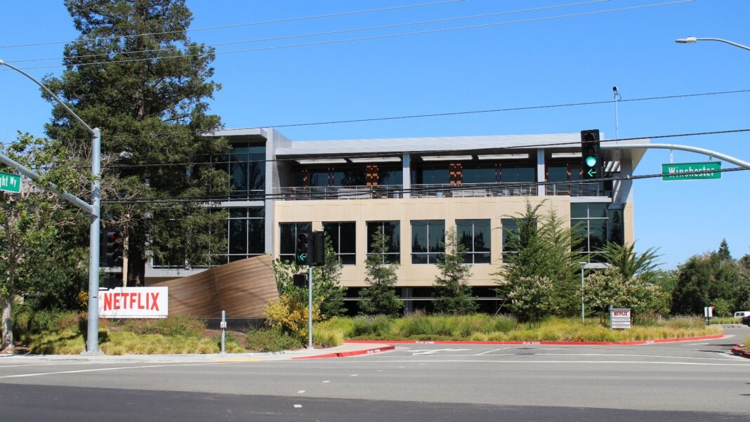 Netflix Cuts About 1,000 Jobs, or 6% of Staff, With Product, Tech, Legal and HR Hit Hardest A two-story modern office building with large windows behind a landscaped corner, a red Netflix sign at the left and traffic signals over an empty intersection under a clear blue sky.