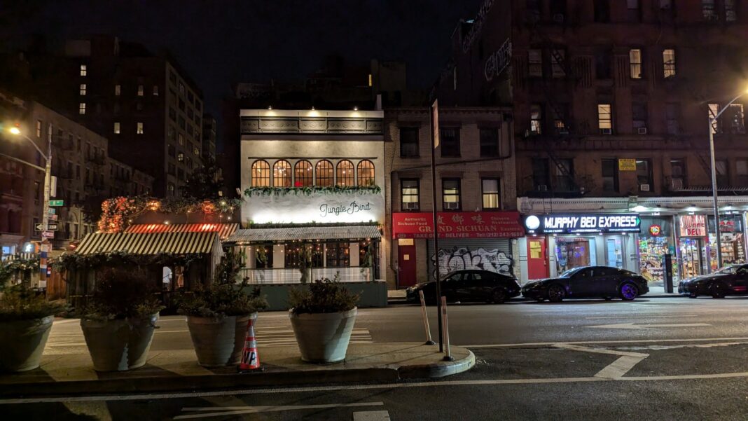 A nighttime street corner with a restaurant's striped awning and planters, lit storefronts including a mattress shop, and parked cars along a wide avenue.