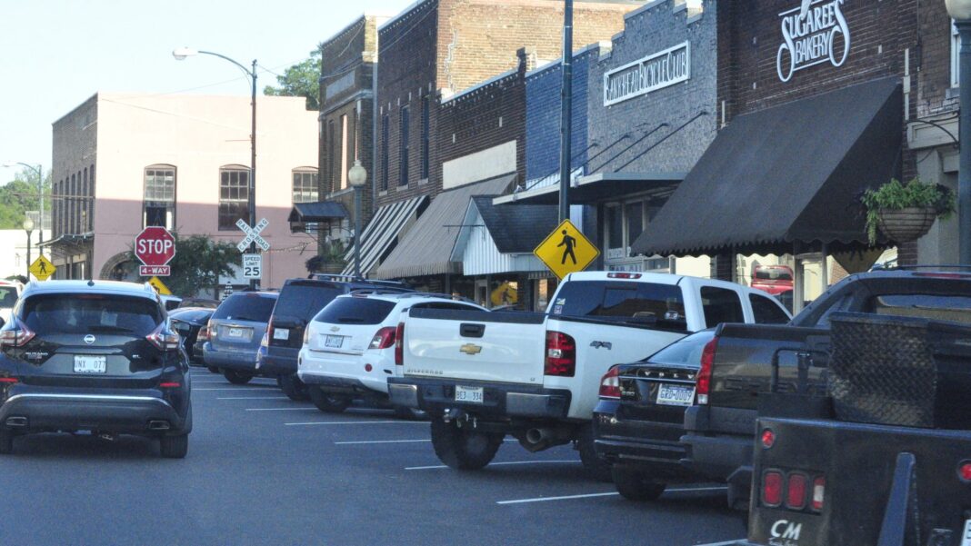 Brick storefronts with awnings along a small-town main street, with pickup trucks and cars parked in the middle of the street and a pedestrian crossing sign