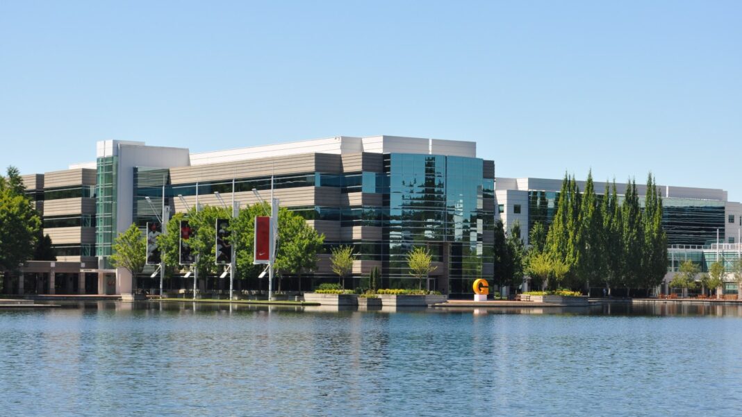 Nike Plans More Job Cuts as It Reshapes Its Global Operating Model Glass-and-steel office buildings with red banners on the far side of a lake on Nike’s corporate campus