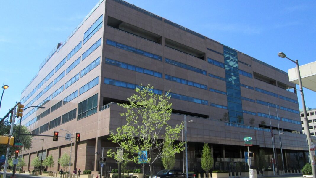 Philadelphia Fed Survey: Factory Hiring Gauge Edges Down to 9.1 as Hours and Hiring Plans Cool A large tan multistory office building with bands of blue windows on a sunny city street corner, with traffic lights and a young tree in front.