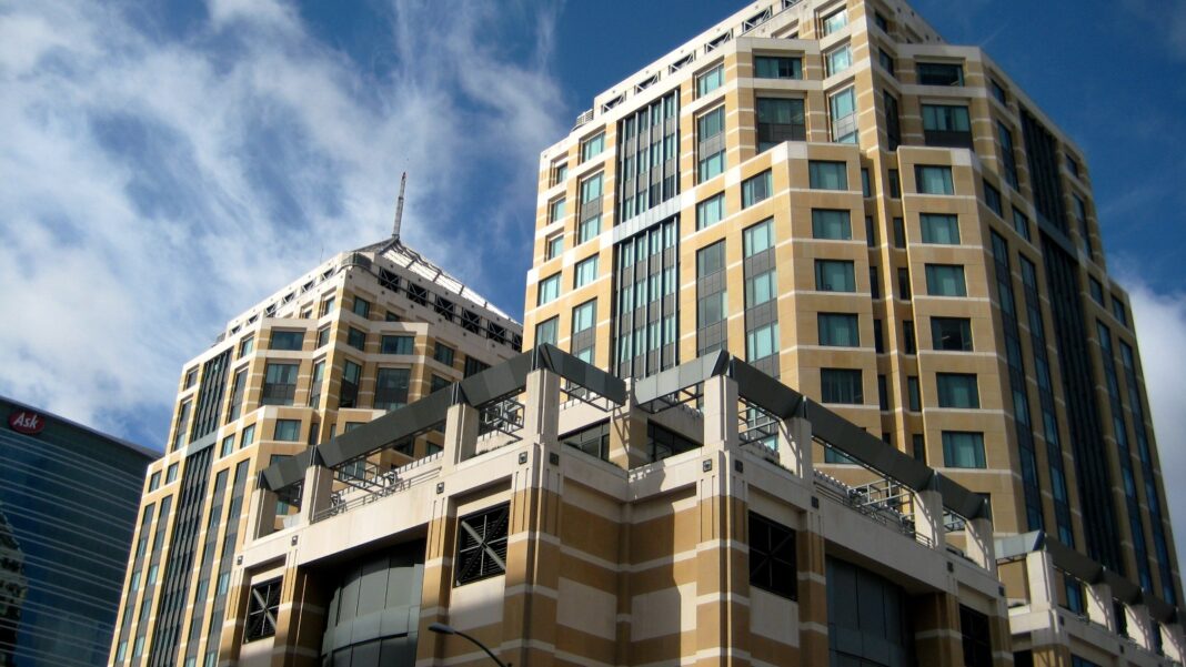 Two tall beige and tan office towers with copper-colored rooftop structures under a blue sky with wispy clouds