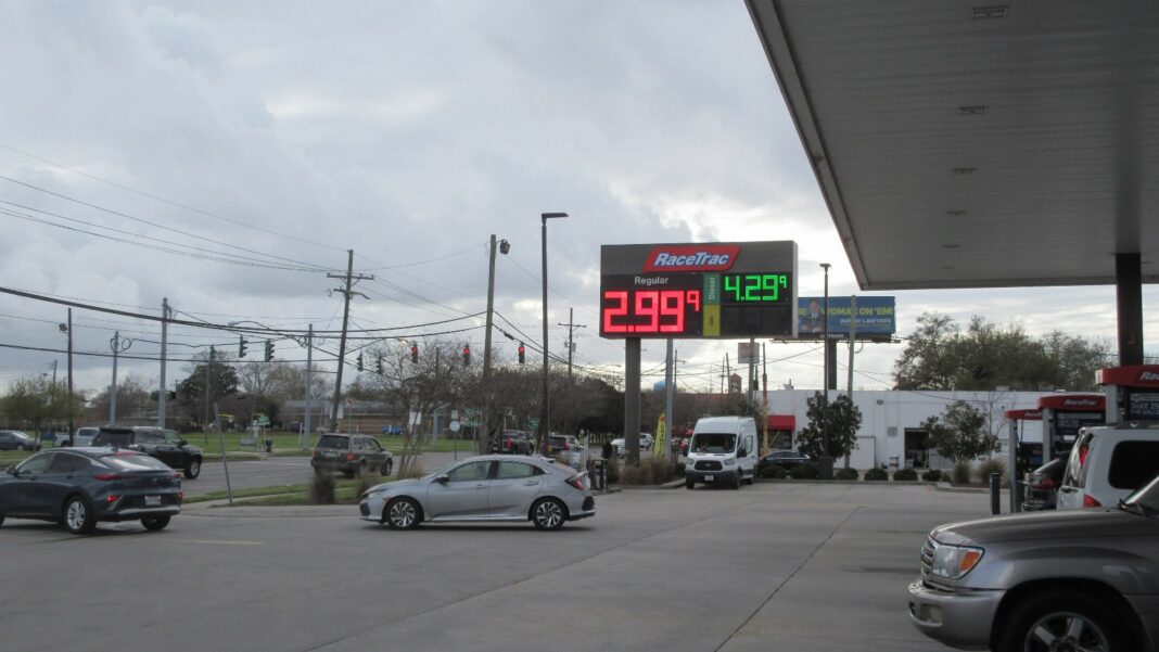 A gas station forecourt under an overcast sky, with a tall red-and-green RaceTrac sign showing digital fuel prices and several cars parked or passing nearby.