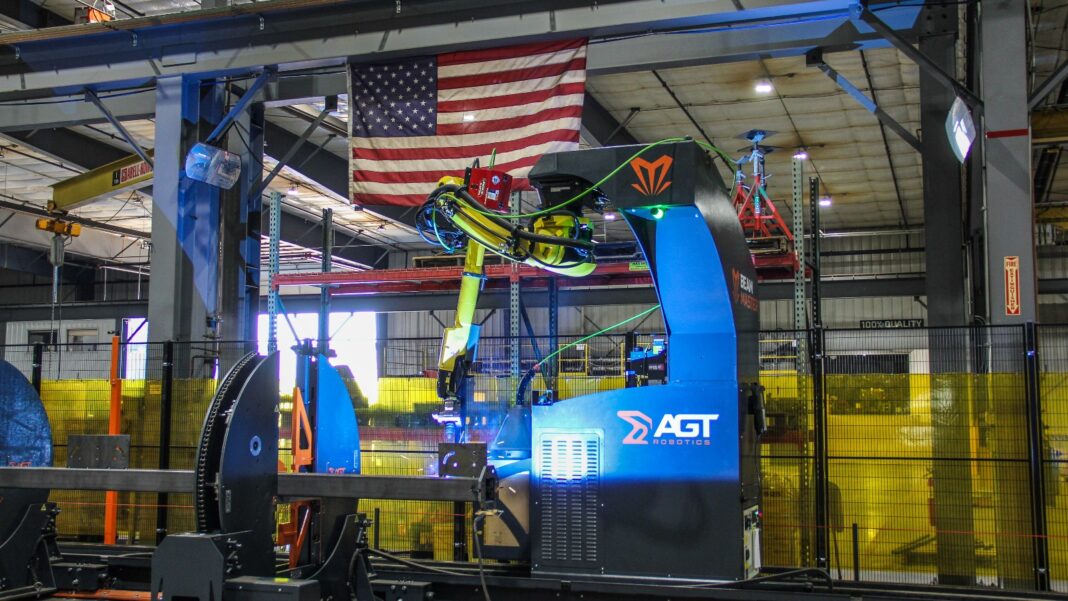 Factory Output Rises 0.3% in September as Utilities Lift Industrial Production A yellow robotic welding arm mounted on a blue machine housing inside a factory bay, surrounded by yellow safety screens and steel framing, with an American flag hanging from the ceiling.