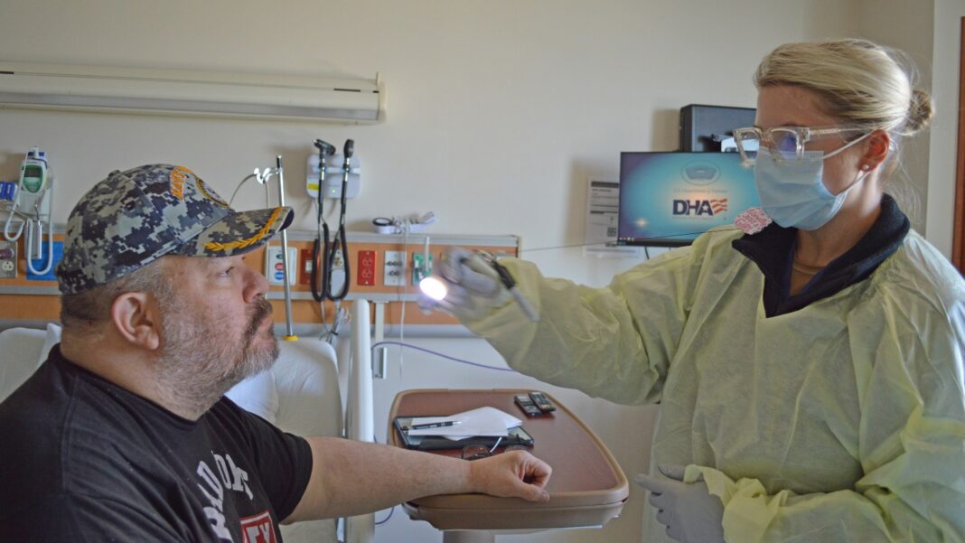 A nurse in a yellow protective gown, mask and glasses shines a penlight at the eyes of a seated patient in a hospital room