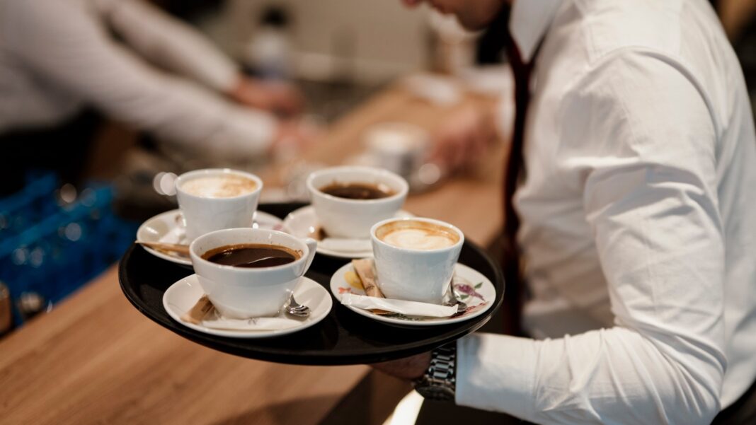 Services Hiring Returns to Growth After Two Months of Contraction A server in a white shirt carries a round black tray holding several cups of coffee past a wooden café counter