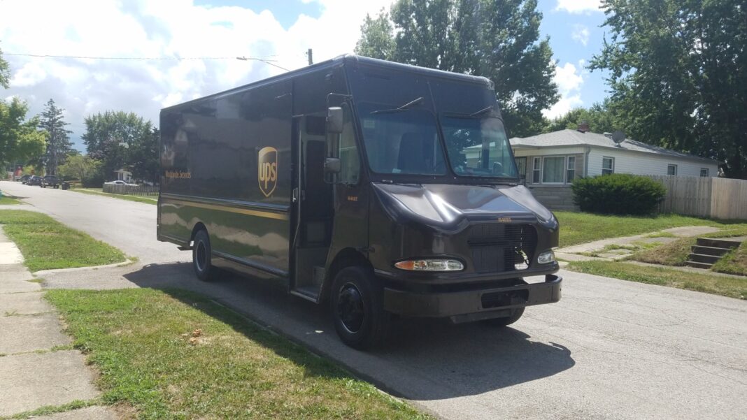 A brown UPS delivery truck with the company shield logo parked on a residential street beside a lawn and trees