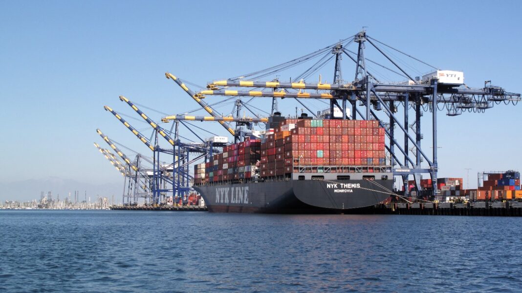 U.S. Import Prices Rise 1.1% in September, Biggest Annual Gain Since 2022 A black-hulled container ship stacked with red and gray shipping containers sits at a dock beneath a row of tall blue and yellow container cranes, with calm blue water in the foreground.